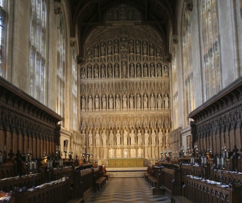 A chapel with a large stone reredos with many statues. The floor is black and white marble and there are wooden pews to either side.
