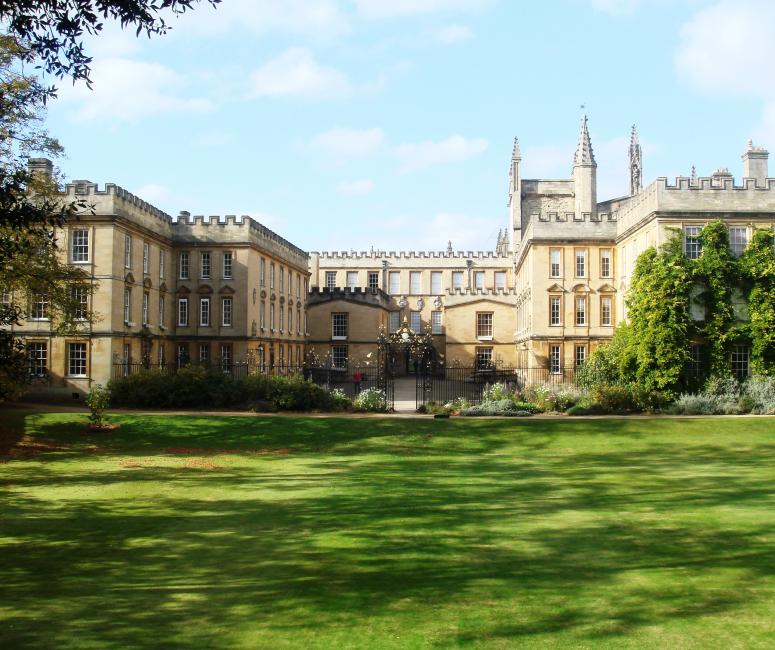 A large stone building with a courtyard and gate leading out onto a grassy garden.