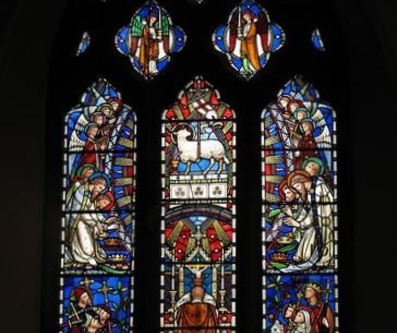 A stained glass window with a lamb, angels and saints, lit by sunlight in a dark church interior.
