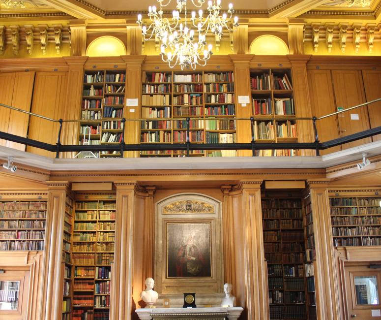 A library with tall bookcases, a balcony, an oil portrait of a man and a chandelier.