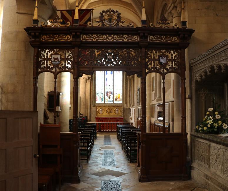 A carved wooden screen in a stone cathedral building with a clear arched window in the background. A stone tomb is visible to the right-hand side.