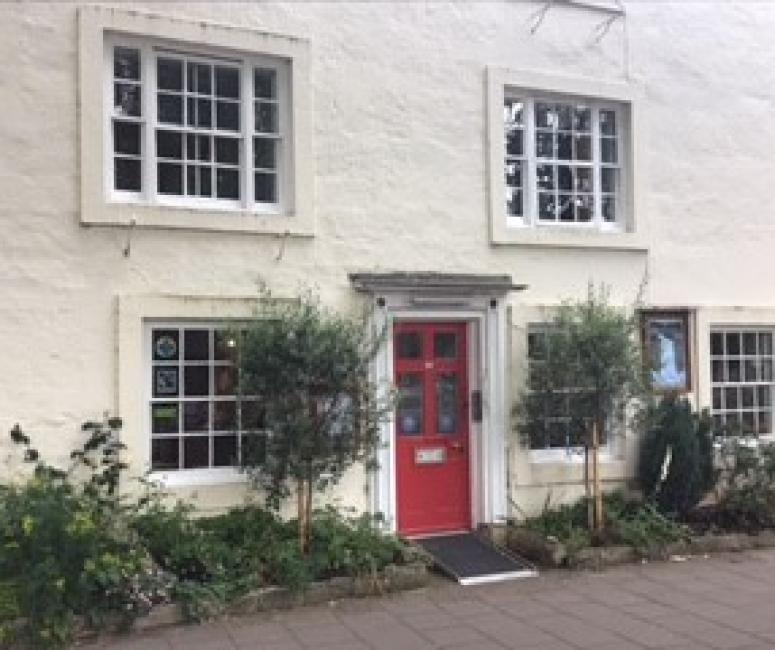 A white building with five sash windows and a red door opening onto the street, framed by two small trees.