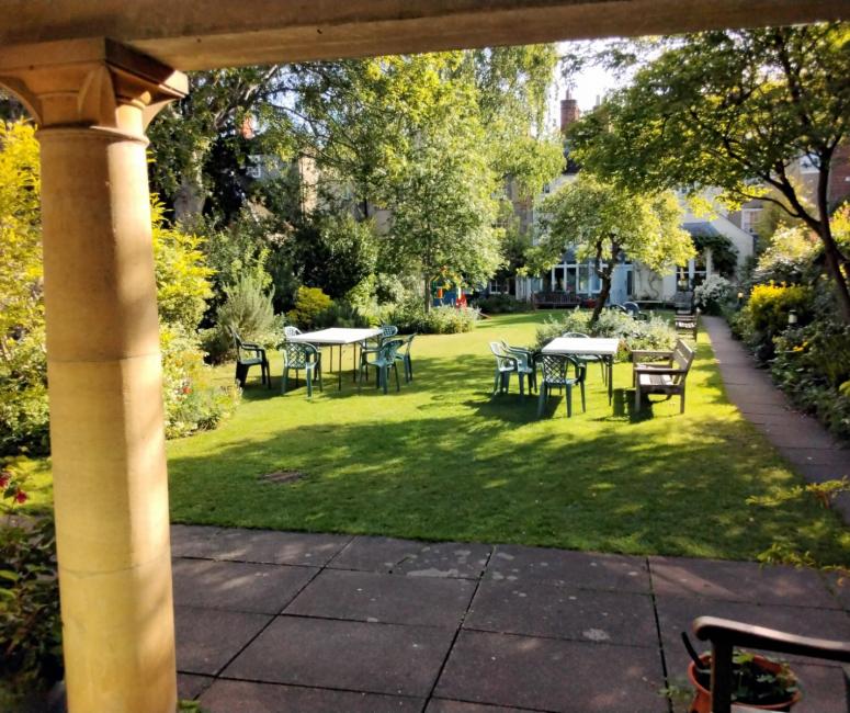 A garden with tables and chairs in dappled shade, viewed from beneath a stone portico.