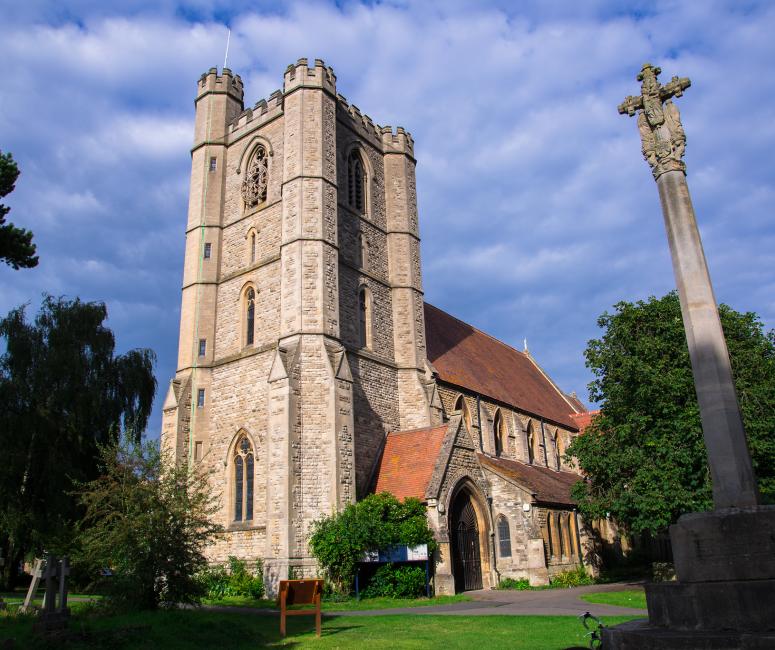 A stone church with a tower and terracotta-coloured roof tiles. A sculpted stone cross on a post is in the foreground.