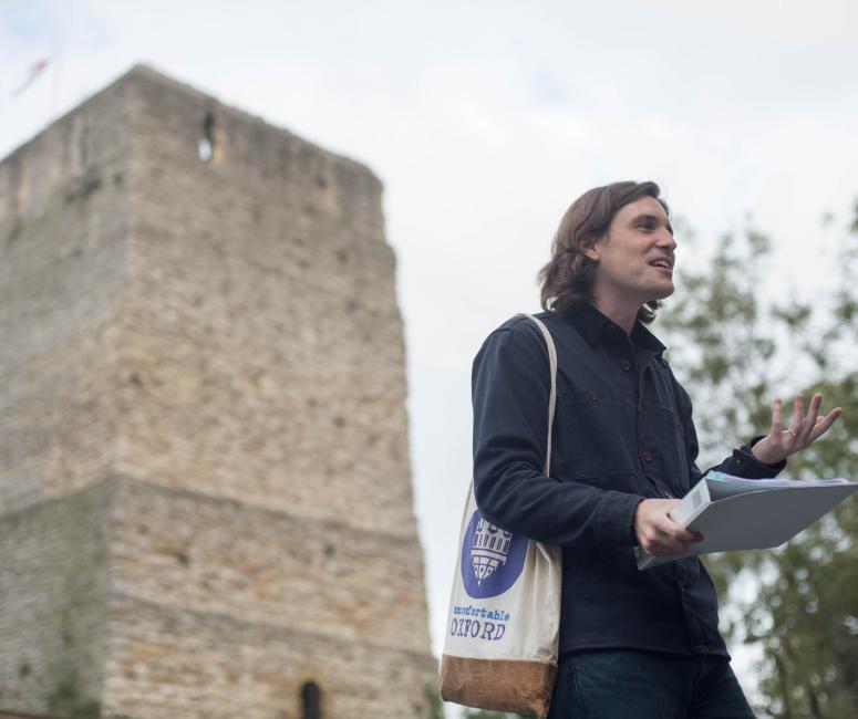 A tour guide with a tote bag that says Uncomfortable Oxford speaking in front of a square stone castle tower.