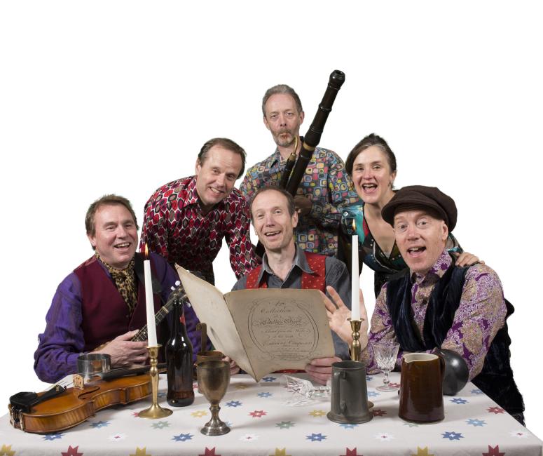 A group of musicians gather around a table with tankards, a candlestick and sheet music.