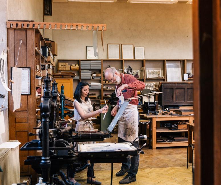 A man helps a woman operate a historic printing press in a workshop with wooden shelves.
