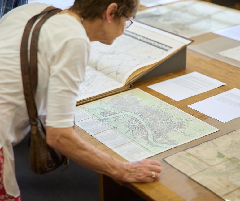 A women leans over a table to look at a series of historic maps on display.