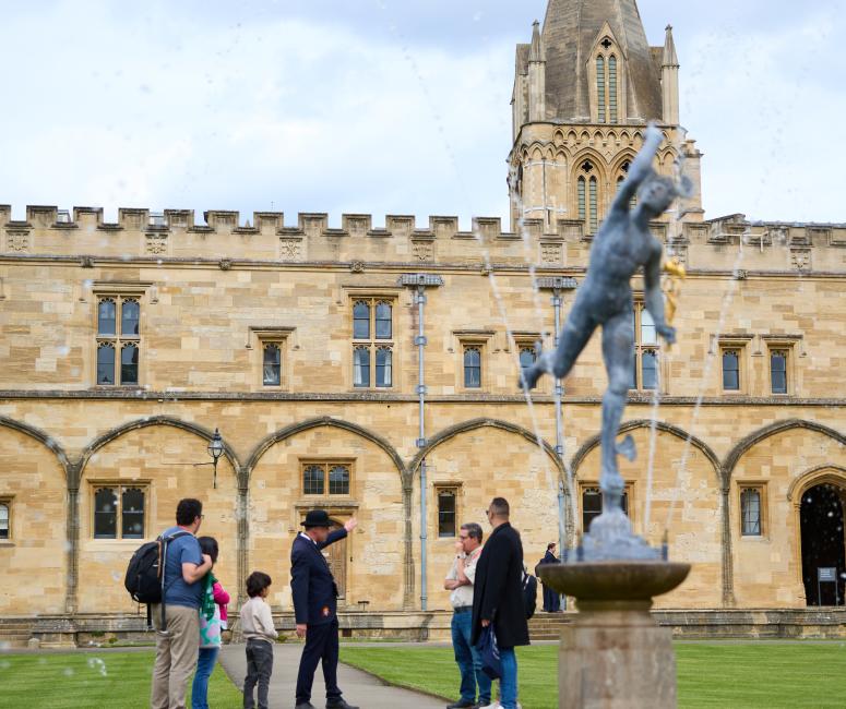 A group of visitors look around in a quadrangle surrounded by stone buildings, with a fountain in the centre.