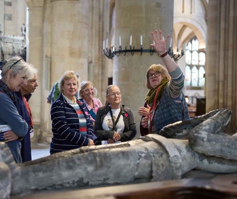 A guide gives a tour to a group in a Cathedral, with a stone effigy monument in the foreground.