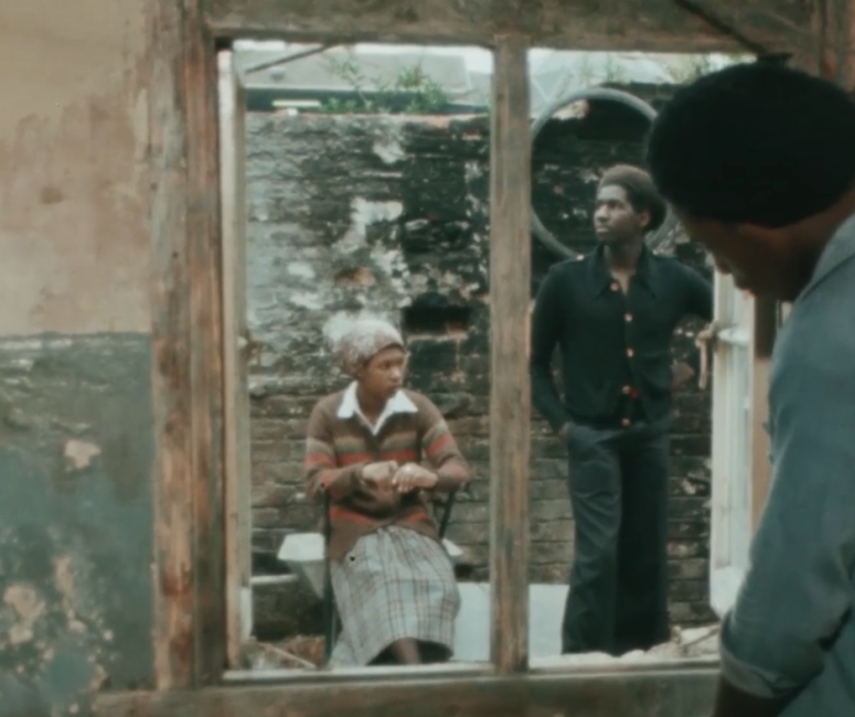 A woman sits and a man stands behind a wooden window frame.