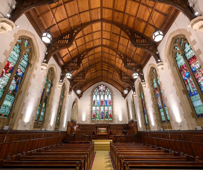 A chapel interior with wooden roof, large arched stained glass windows and dark wooden pews.