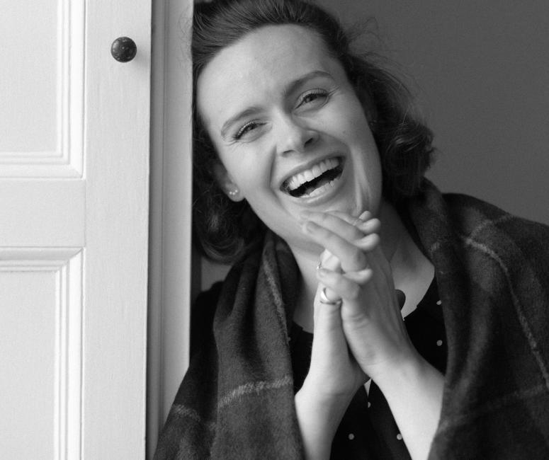 A black and white photograph of a woman laughing. Her hands are clasped together and she is leaning against a wooden panel containing a door.