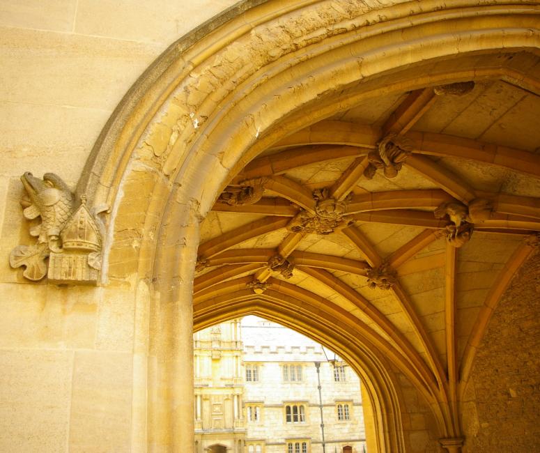 The archway of a medieval gatehouse with a vaulted ceiling and a stone college building beyond.
