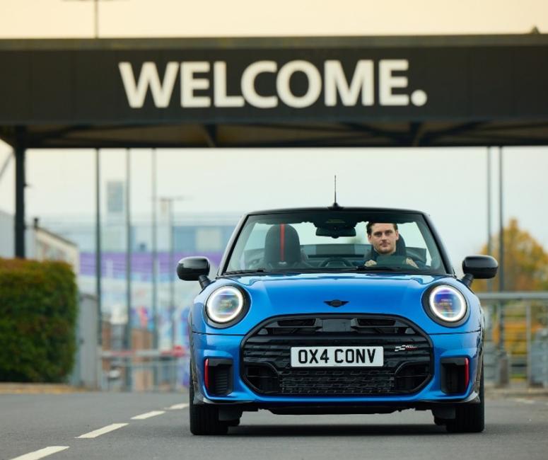 A blue Mini drives towards the camera beneath a black sign with Welcome written in white.
