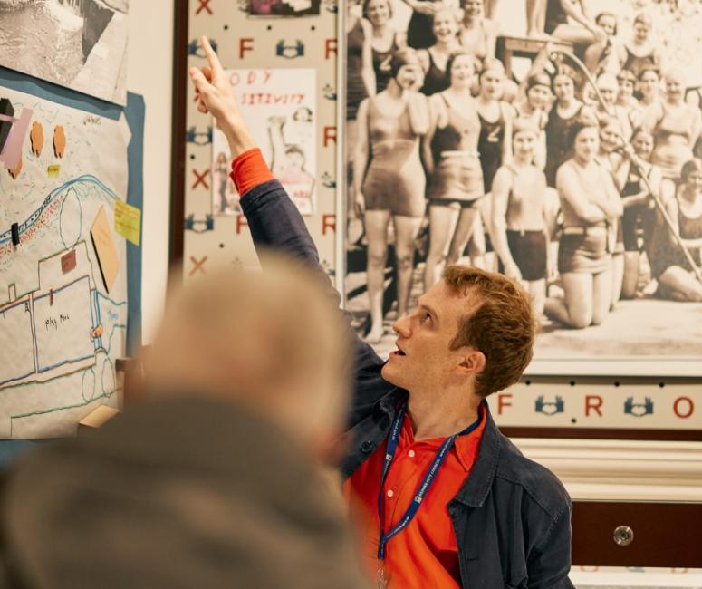 A guide in an orange shirt points at a history display during a tour to members of the public.