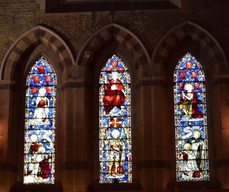 A row of three stained glass windows depicting figures in a dark church interior.