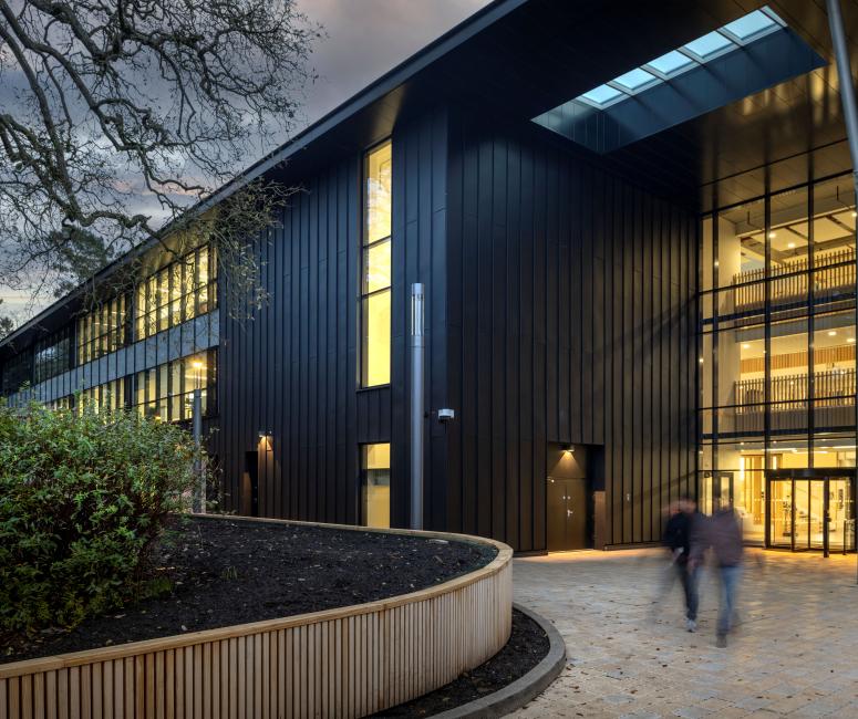 A black modern building with a large full height window and sweeping landscaping.