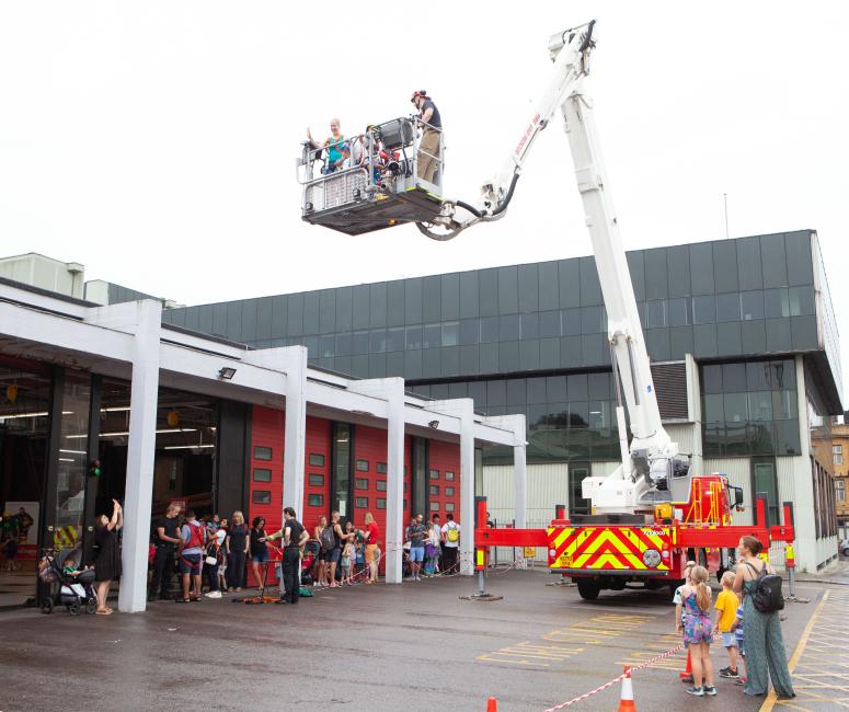 A fire truck with a crane lifting two people. The truck is parked in front of a fire station with red doors.