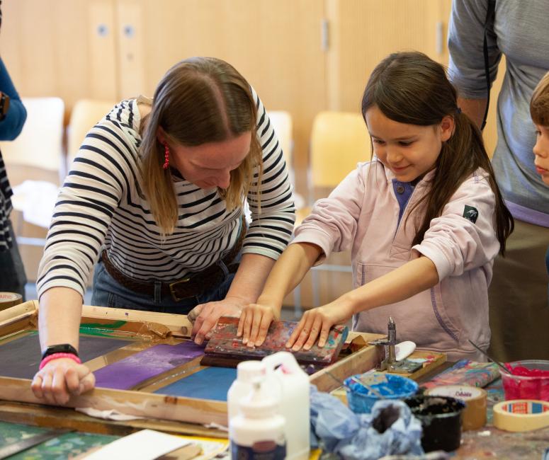 A woman in a stripey top held a girl scrape excess paint off a silk-screen printing frame. They are surrounded by paint pots and a boy looks over the girl's shoulder.