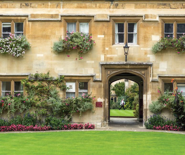 A seventeenth-century stone building with window boxes and a lush border and a passage through to another quad.