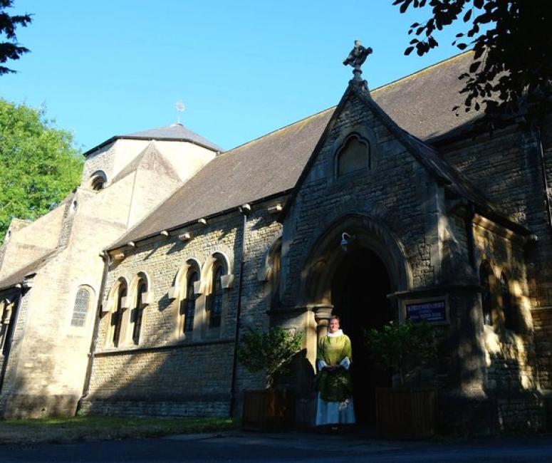 On a sunny day, a shadow is cast across the porch of a stone church, with the rest of the building stretching away into a sunny churchyard.