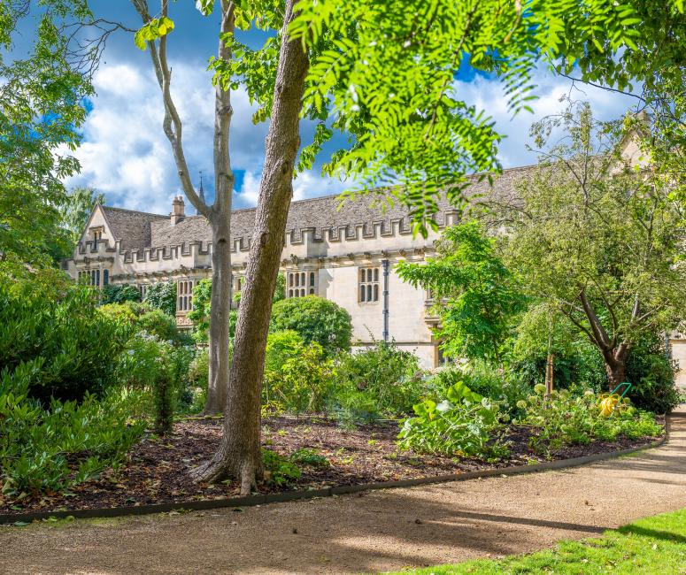 A sunlight garden with trees and shrubs with a crenellated stone building in the rear.