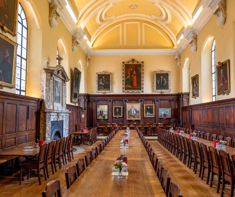 A large dining hall with portraits and panelling on the walls and three long wooden tables running the length of the room.