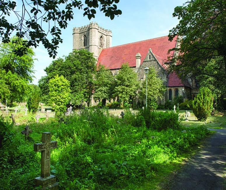 A lush churchyard with stone headstone and a path leading to a red roofed stone church with a tower.