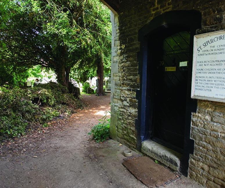 A path leading around a stone church building into a shaded tree-filled cemetery beyond.