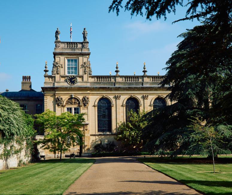 A wide path through a grassy area, leading to a seventeenth-century stone building with a tower, windows with rounded arches and an arched passageway through the building.
