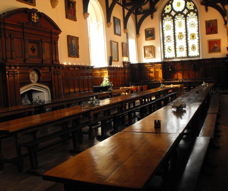 A dining hall with long wooden tables and benches, panelling covering the lower half of the walls with portraits displayed above, beneath the wooden beams of the ceiling.