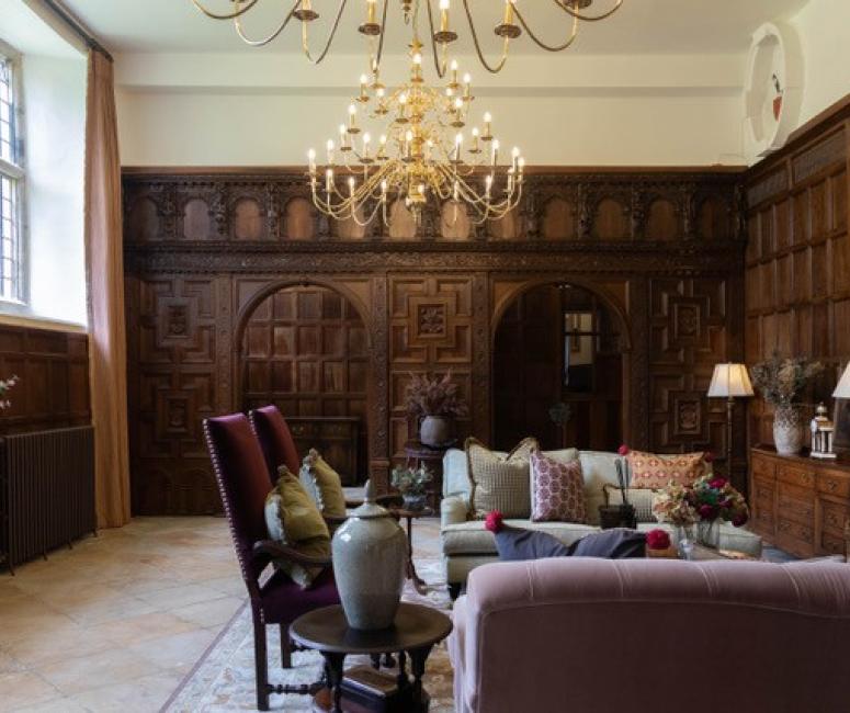 The interior of a seventeenth-century manor house with dark wooden panelling, a large chandelier and large windows with many small panes of glass.
