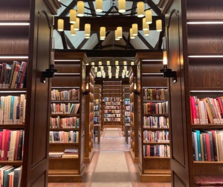 A library interior with tall bookcases and ironwork chandeliers. The roof is held up by wooden beams.