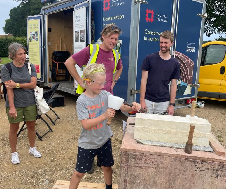 A boy has a go at stone carving with a chisel and mallet, with three adults standing watching next to an English Heritage Conservation in Action van.