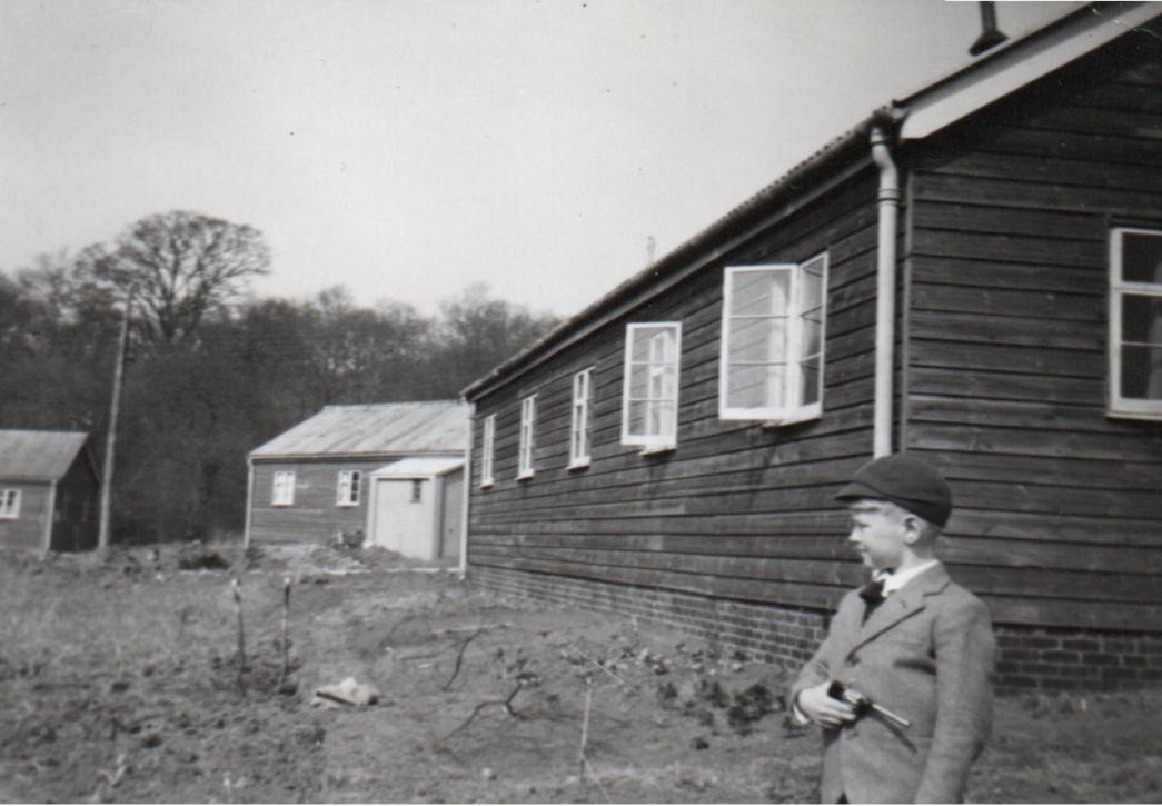 A black and white photograph of a child standing in front of wooden hut in a grassy area.