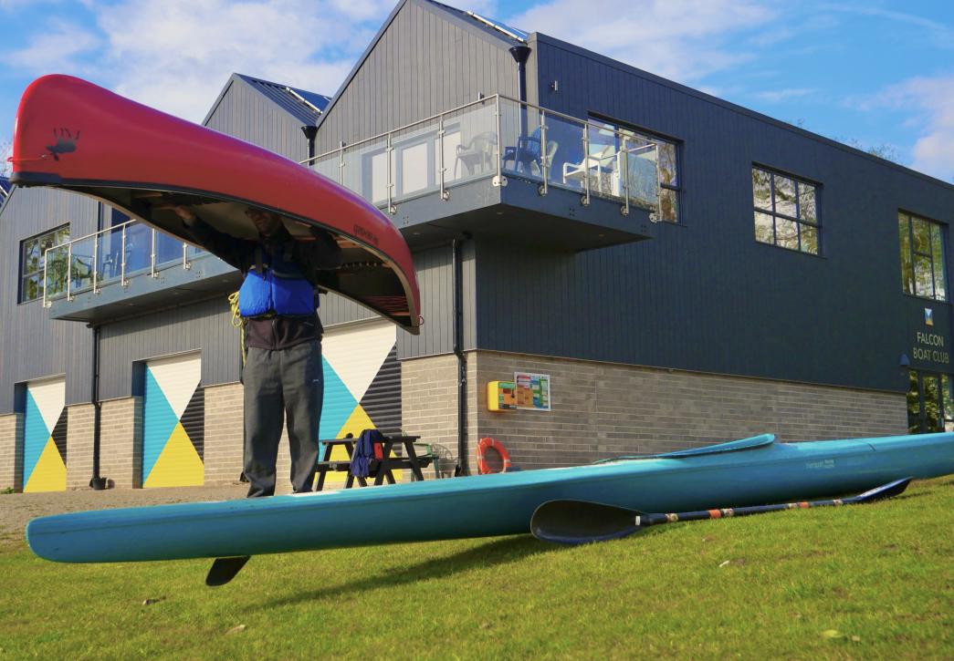 A photograph of a grey boathouse with large doors painted blue, white, black and yellow. In the foreground a man lifts a red canoe over his head. A blue kayak sits on the grass.