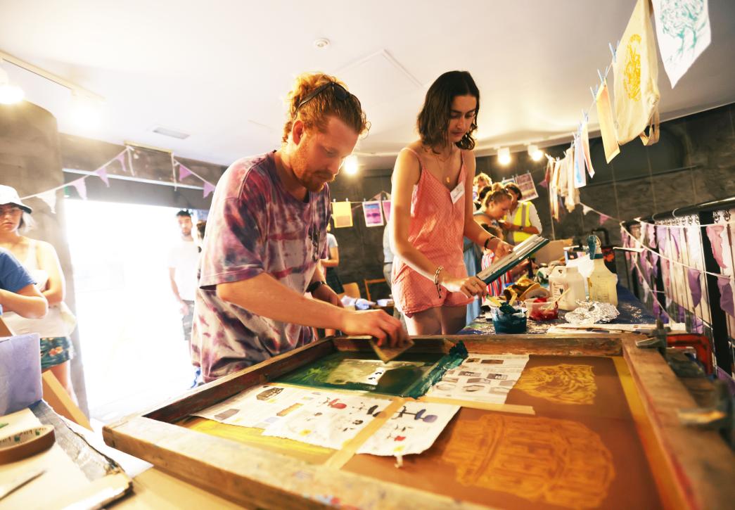 Two people leaning over a wooden printing frame with prints drying on pegs on a line above.