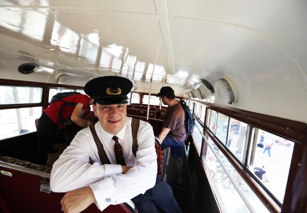 A man wearing a hat that says inspector looks at the camera inside a vintage bus with a white ceiling.